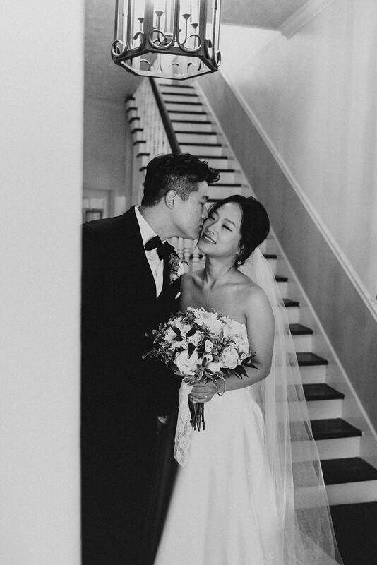 black and white photo of groom kissing bride's cheek in farmhouse wedding venue foyer