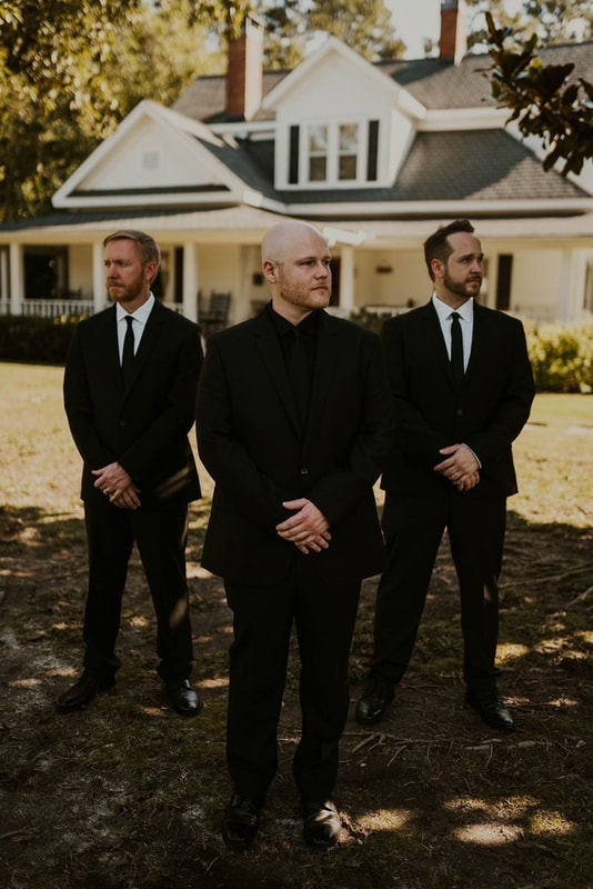 groom in all black with groomsmen in black suits posing in front of farmhouse