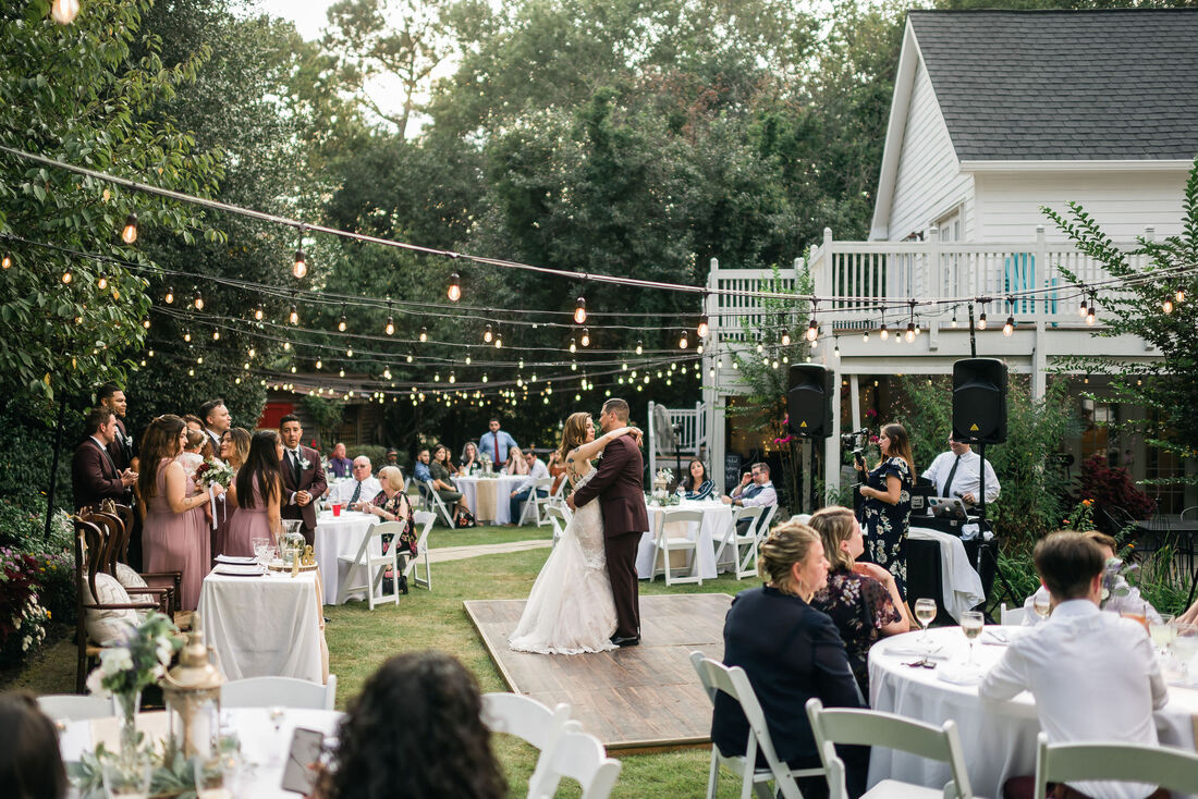 Couple's first dance on outdoor dance floor under string lights while guests and bridal party watch.