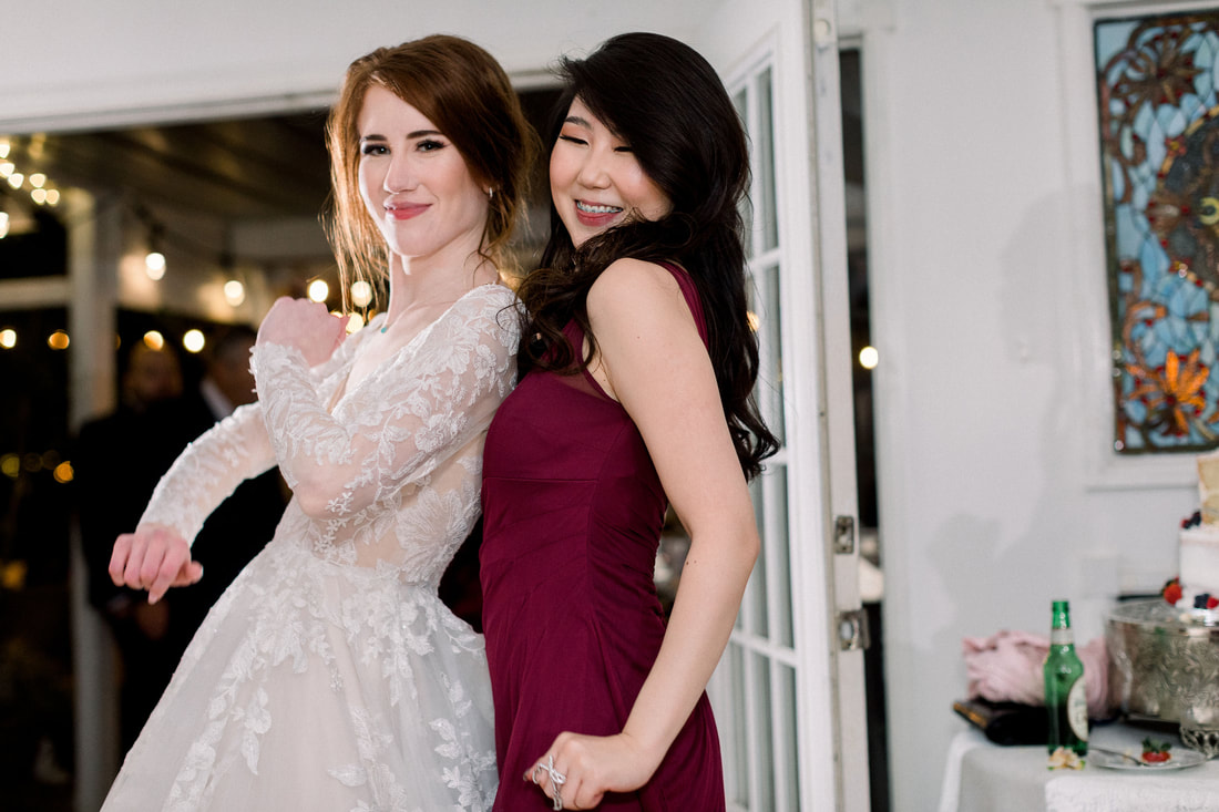 Bride in lacey, long-sleeve dancing in carriage house with bridesmaid in deep burgundy dress.
