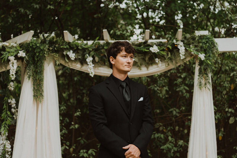 groom in all black standing at garden altar