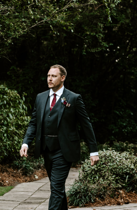groom in charcoal suit with burgundy details walking through garden path to ceremony