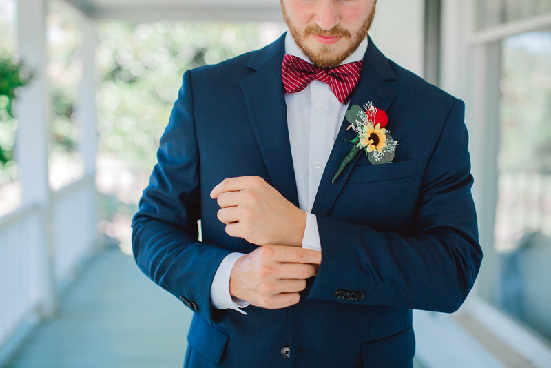 groom adjusts sleeves of navy suit while wearing red striped bow tie and boutonniere with a small sunflower and red rose