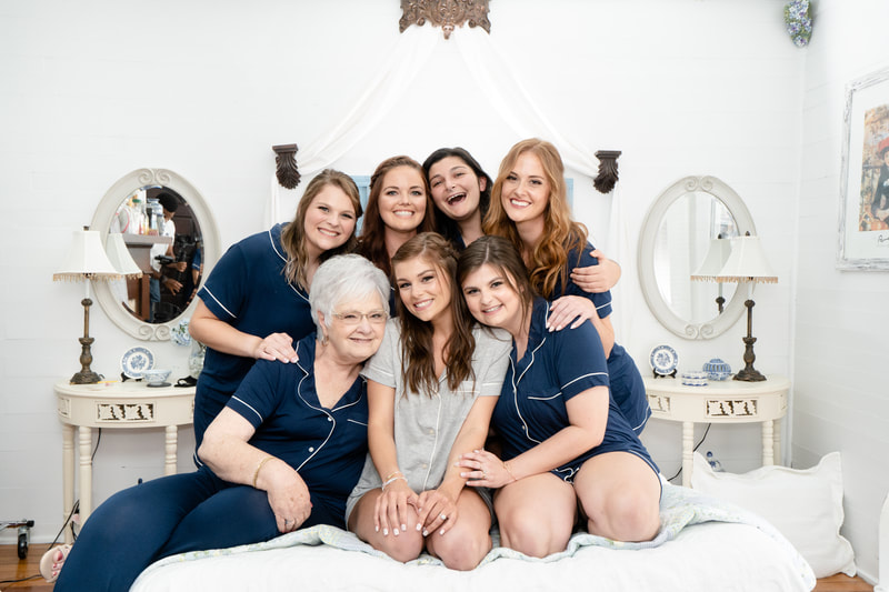 bride in grey outfit and bridesmaids in navy getting-ready outfits sitting on farmhouse bed