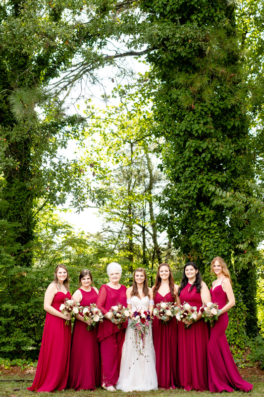 bride standing in middle of bridesmaids in burgundy dresses surrounded by ivy-covered trees