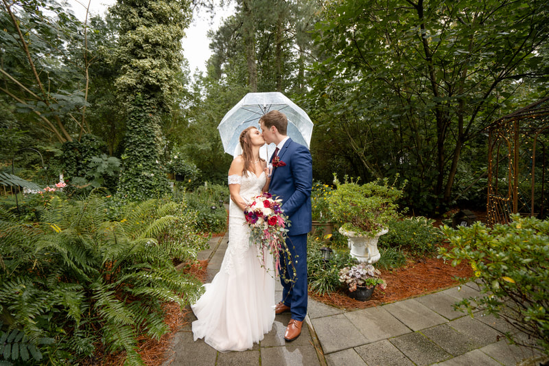 newlyweds kissing under clear umbrella as it rains in Four Oaks' gardens