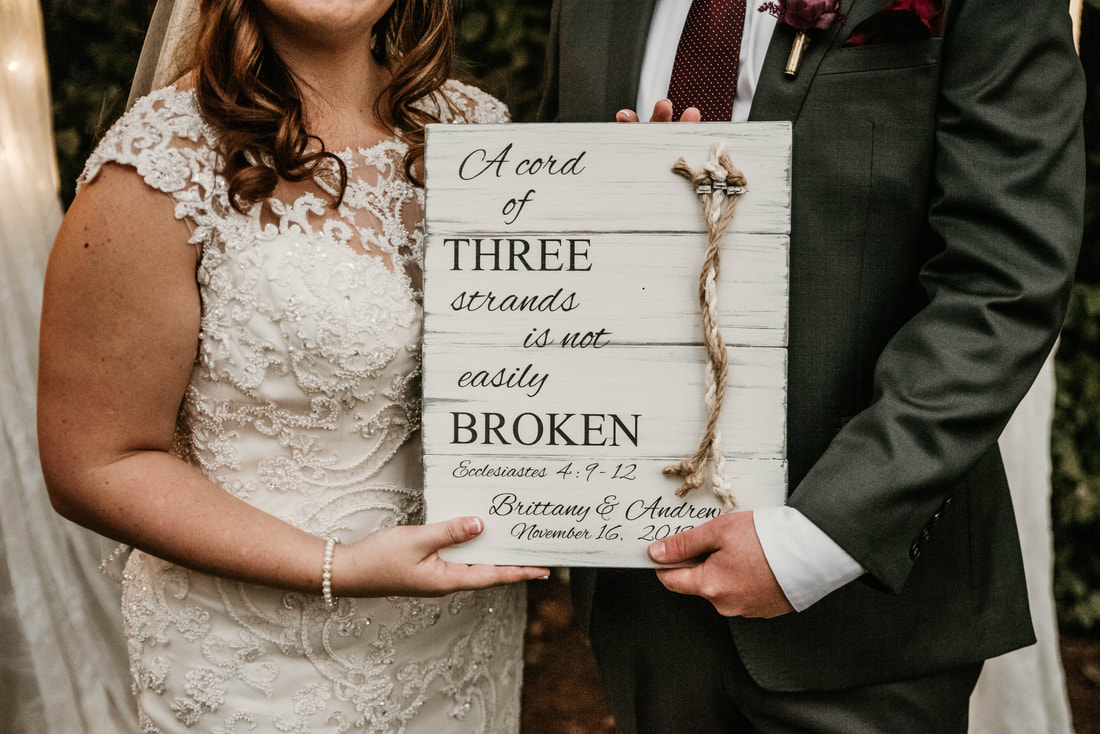 Brittany and Andrew holding white wood plaque with three strands of rope and ecclesiastes verse for knot tying ceremony