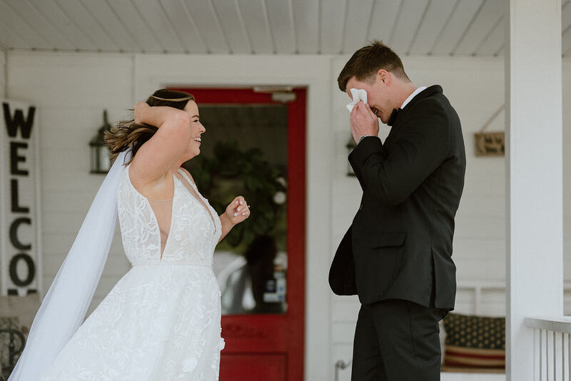 groom wiping tears during first look on farmhouse porch with red door