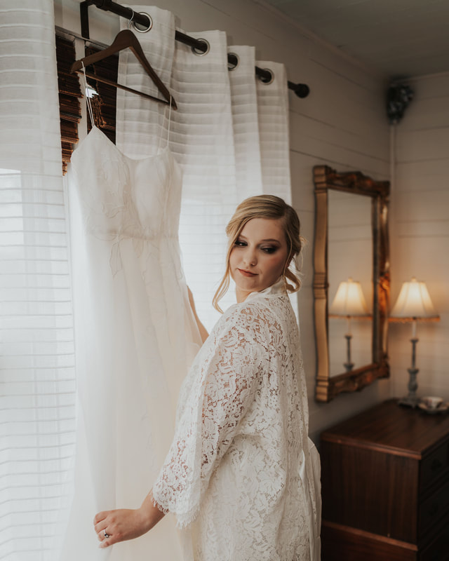 bride in lace robe posing with wedding dress hanging on window