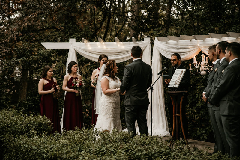 couple with bridal party at outdoor altar surrounded by greenery listening to officiant during ceremony