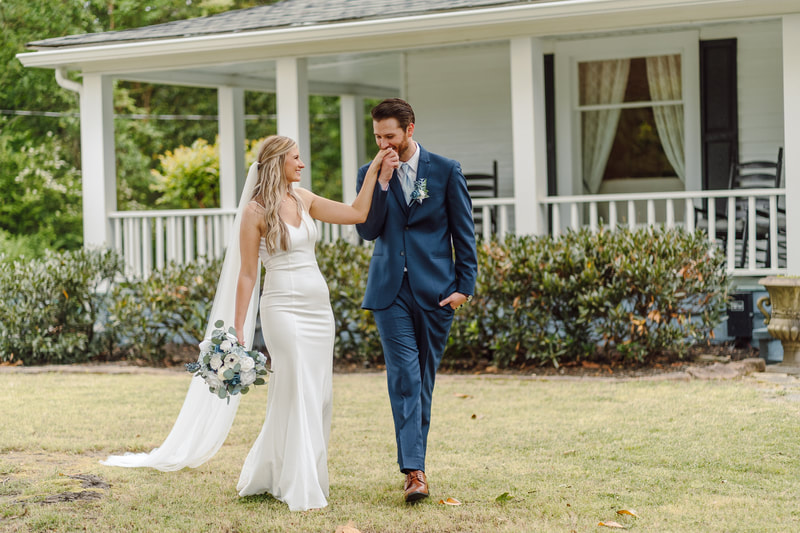 groom kissing bride's hand while walking in front of white farmhouse