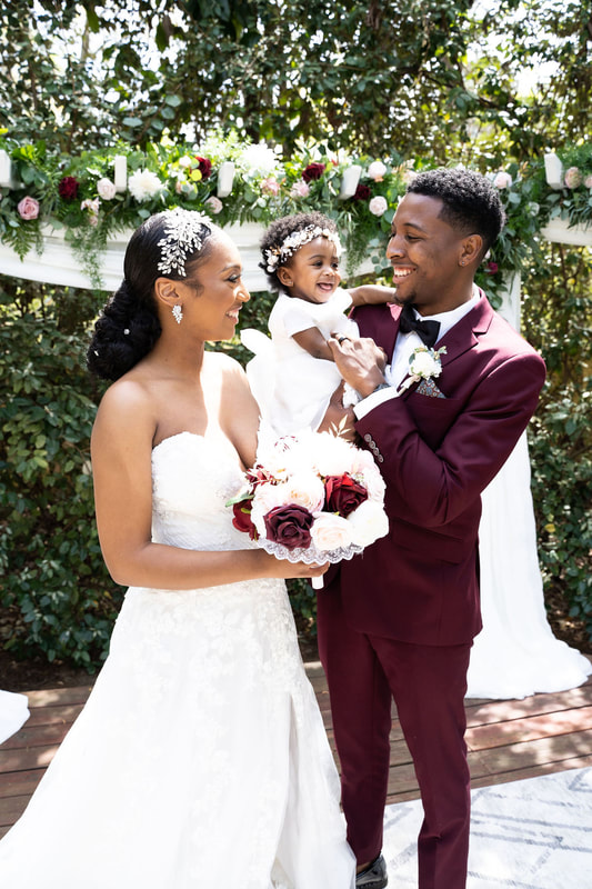 groom in burgundy suit holding baby daughter with bride at garden altar