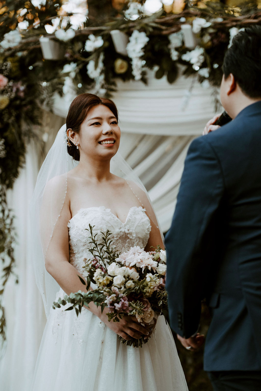 bride smiling at garden altar with greenery and white fabric
