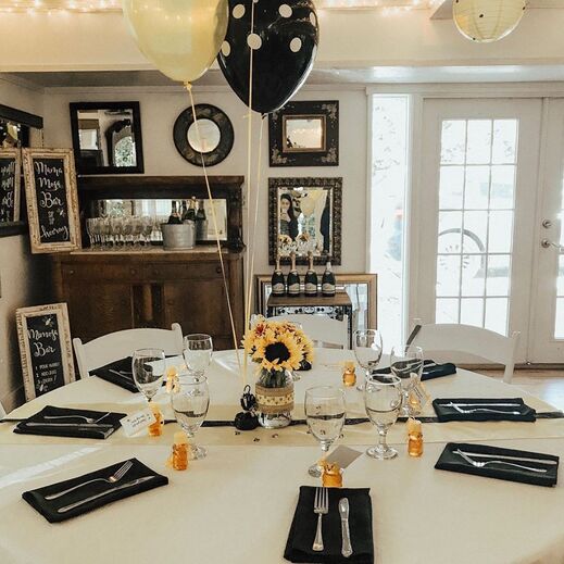 Picture of a bee-themed baby shower, 'Bay-bee.' The round tables are decorated with sunflowers, black napkins, honey favors, sunflower floral centerpieces and yellow and black balloons. A simple mimosa bar is set up in the corner.