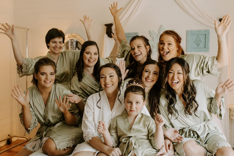 bride in white robe with bridesmaids in sage robes sitting on farmhouse bed cheering