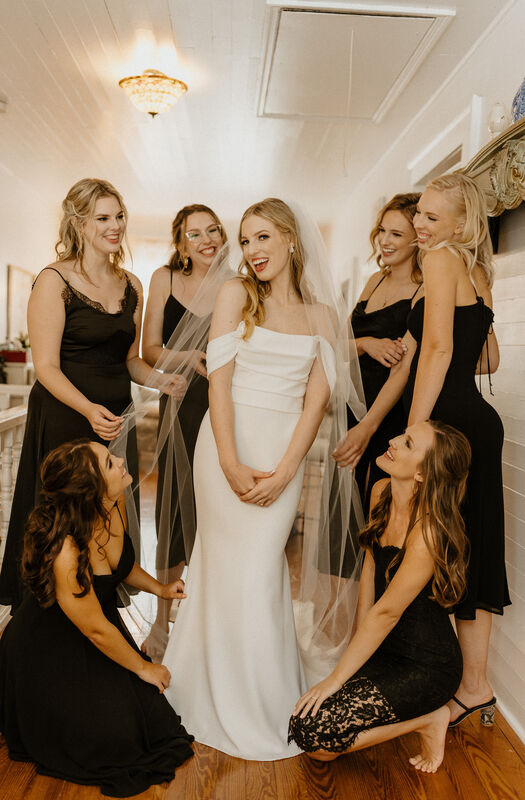 bride in satin dress posing with bridesmaids in mismatched black midi dresses