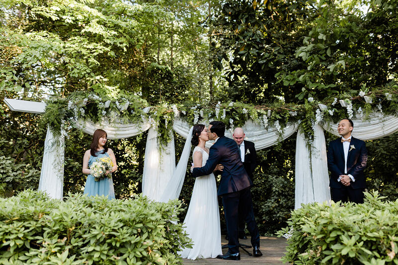 couple kissing at arbor decorated with greenery, and small bridal party