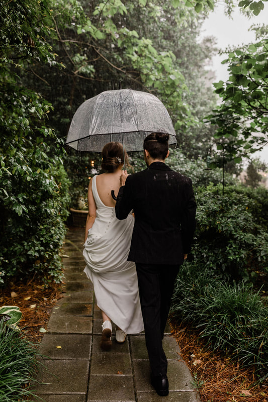 bride and groom walking through garden path under clear umbrella