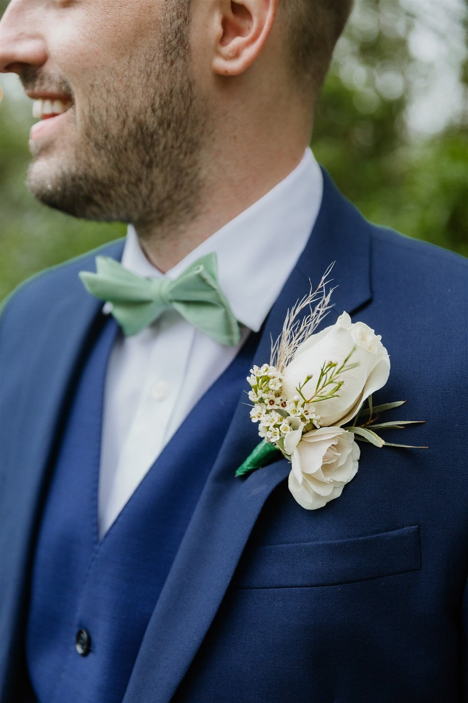 close-up of groom's outfit with navy blue suit, sage green bowtie, and neutral floral boutonniere