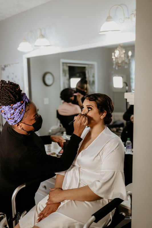 The bride sits in a plush chair in a silk ivory robe while the makeup artist finishes her eye makeup.