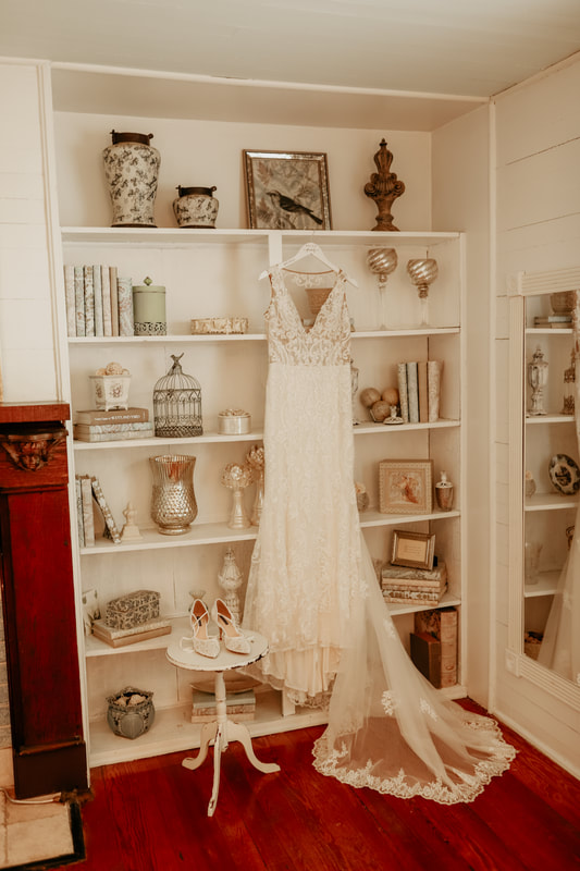 The wedding gown hangs on a white bookshelf filled with knickknacks. The bride's shoes sit on a white table to the left of the dress.