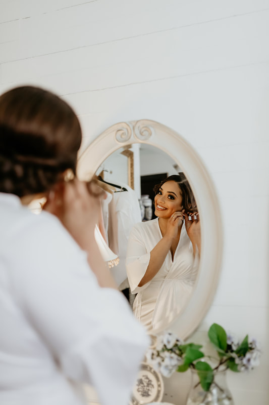 The smiling bride puts in her earring in a mirror as she gets ready in the bridal suite.