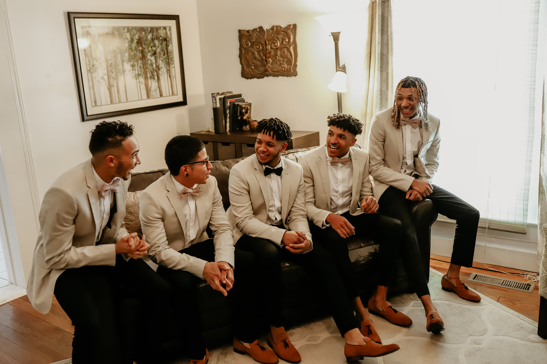 The groom and groomsmen laugh together on a leather couch in the groom's suite.