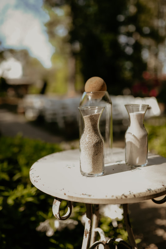 Two glass vases of sand sit on a white table near the edge of the altar area before the ceremony. The white wedding chairs for guests at the ceremony can be seen in the background.