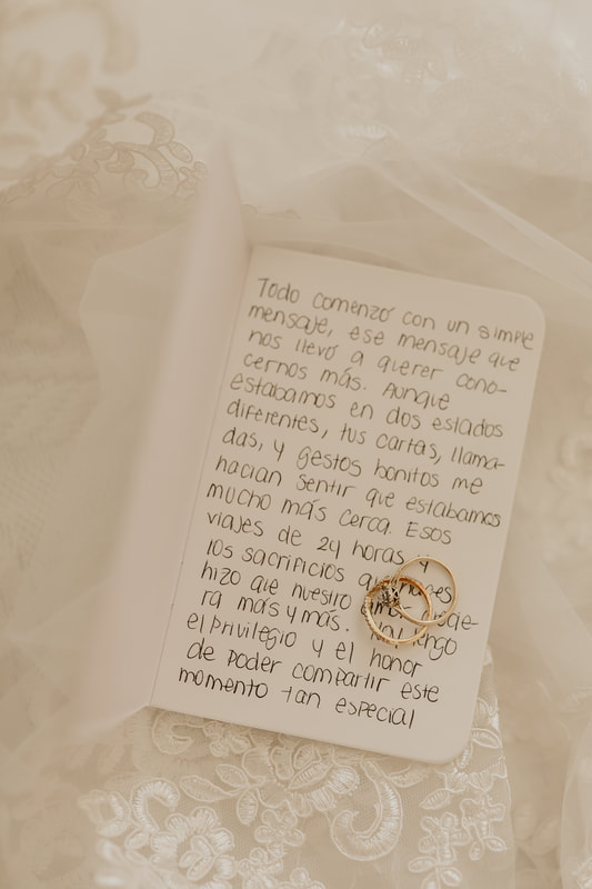 A wedding band and engagement ring sit on top of a card with handwritten vows in Spanish. Lace and tulle from the wedding dress and veil surround it. 