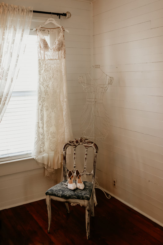 The wedding dress is hung in front of a window while the bride's shoes sit in a white and blue farmhouse chair in front and to the right.