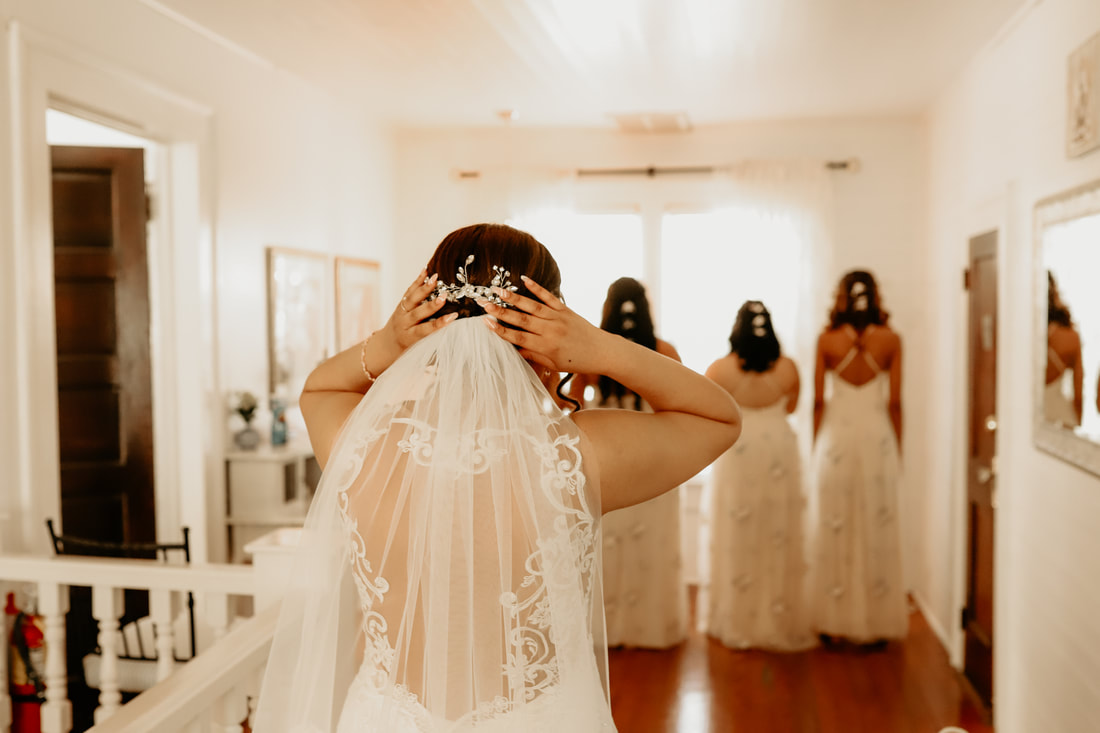 The bride walks towards her bridesmaids while fixing her veil. The bridesmaids are faced away.