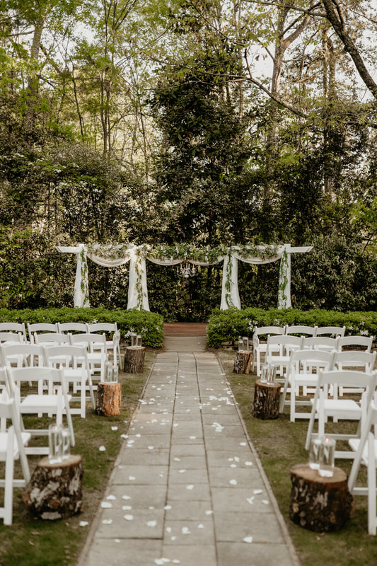 Rows of white wedding chairs line the aisle leading to the altar. Tree stumps with floating tealights in hurricane vases mark every other row, and flower petals are scattered on the aisle. The arbor behind the altar is draped with white chiffon. Full greenery decorates the top, and wisteria hangs down the four posts. A chandelier hangs in the center of the arbor.