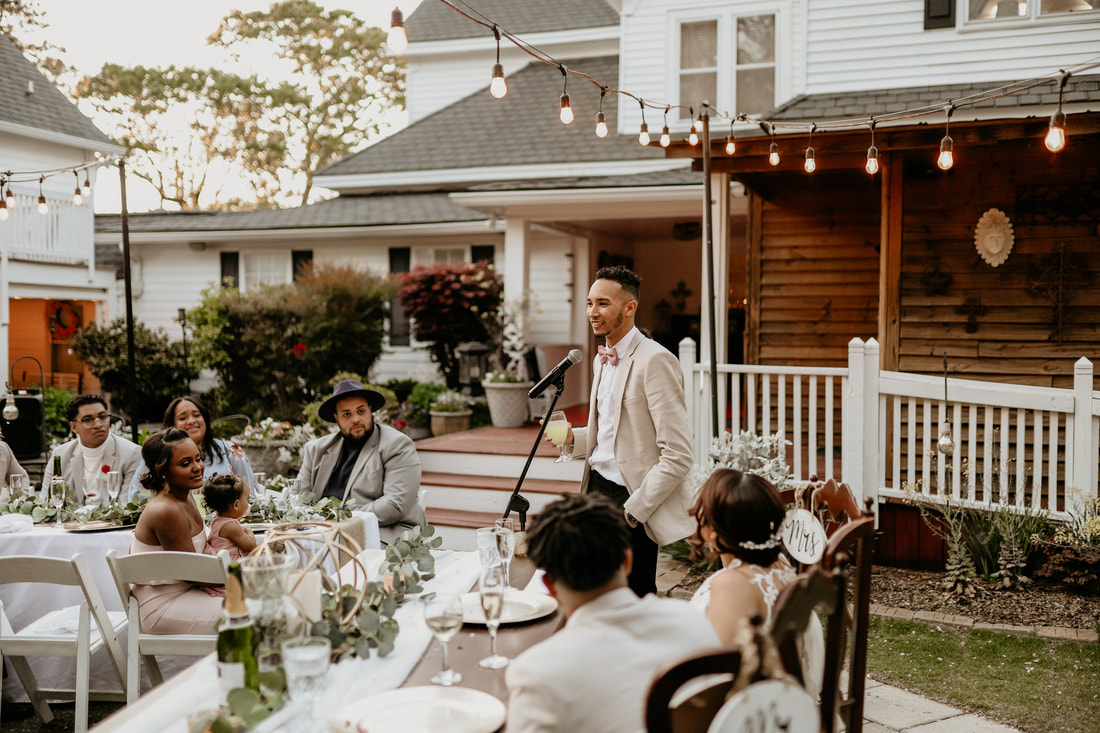 The best man smiles as he gives his speech. The farmhouse is in the background, and the bride and groom are sat at a sweetheart table in front of him.