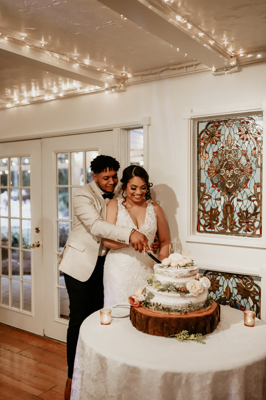 The groom helps his wife cut their wedding cake. The cake is decorated with rustic white buttercream and florals. The cake sits on top of a thick wood slice. Two gold votives with tea lights are on either side of the cake. The round table has a white tablecloth and lace overlay.