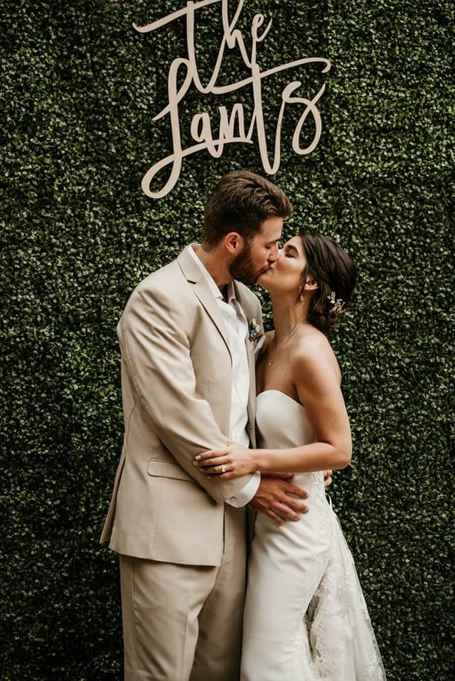 couple sharing kiss in front of greenery wall with custom name sign