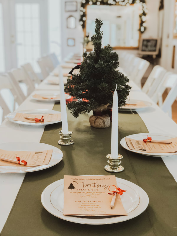 Christmas party or dinner set up. Green runners are decorated with gold candlesticks and mini Christmas trees. Place settings are simple gold or silver rimmed china with parchment menus on top.
