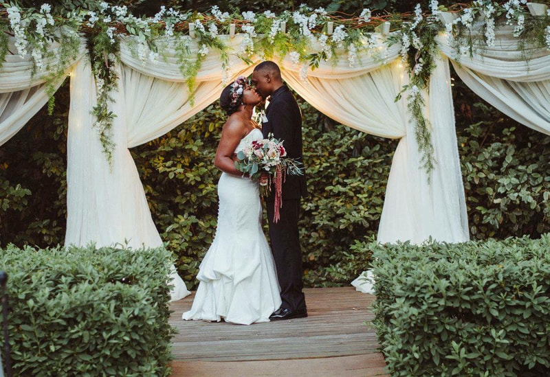 newlyweds kissing under ceremony altar with white chiffon, greenery and white flowers