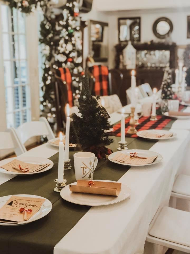 Christmas dinner banquet table set up. Gold candlesticks, mini Christmas trees, and flannel accents on dark green runners. Parchment menus with key favors sit on top. Christmas mugs are used as glasses.