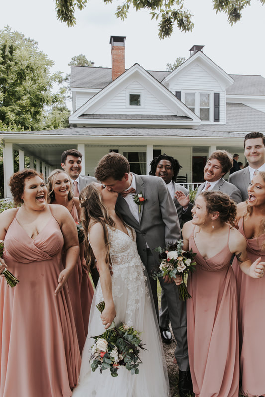wedding party around bride and groom kissing in front of farmhouse