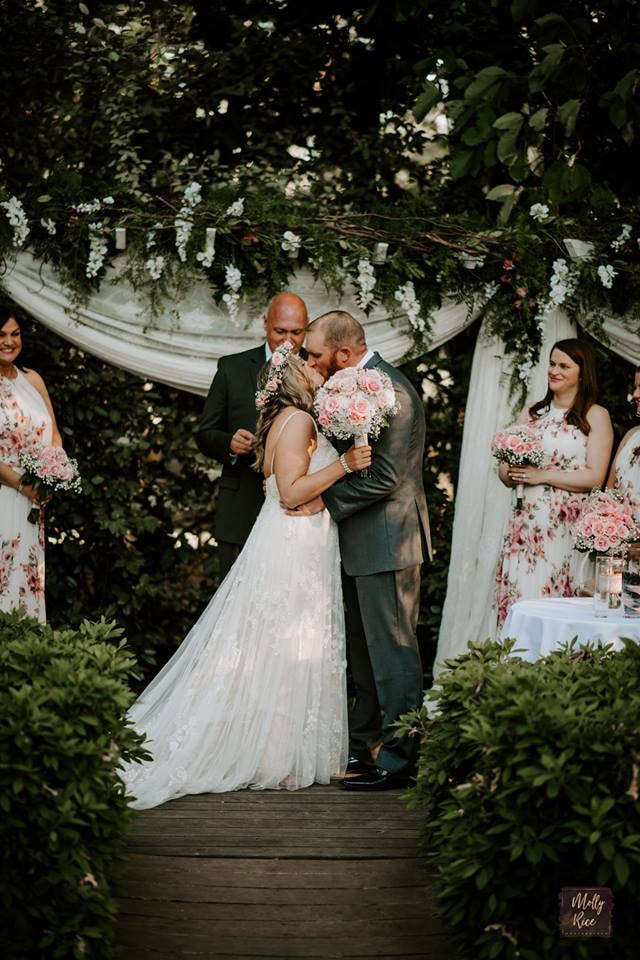 bride and groom's first kiss by altar covered in greenery and white flowers