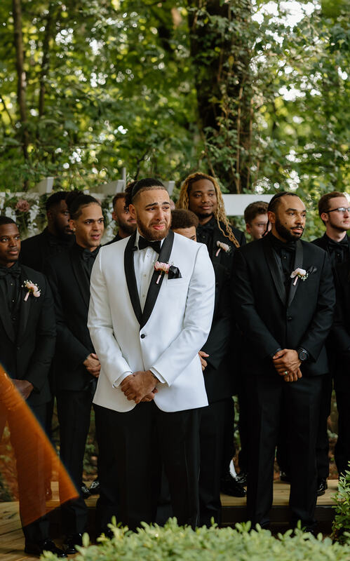 groom in white tux crying at altar with groomsmen in all-black behind him