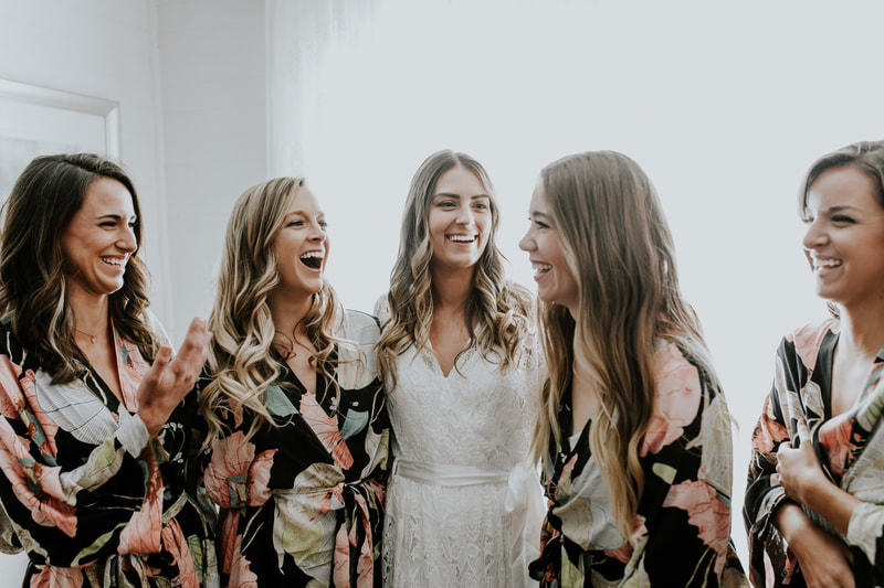 bridesmaids in floral robes laughing with bride in white lace robe
