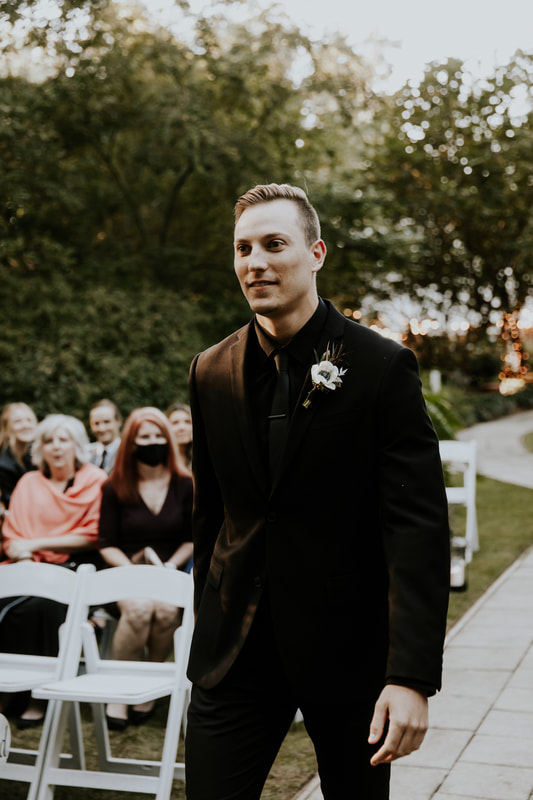 groom in all-black suit walks down outdoor ceremony aisle