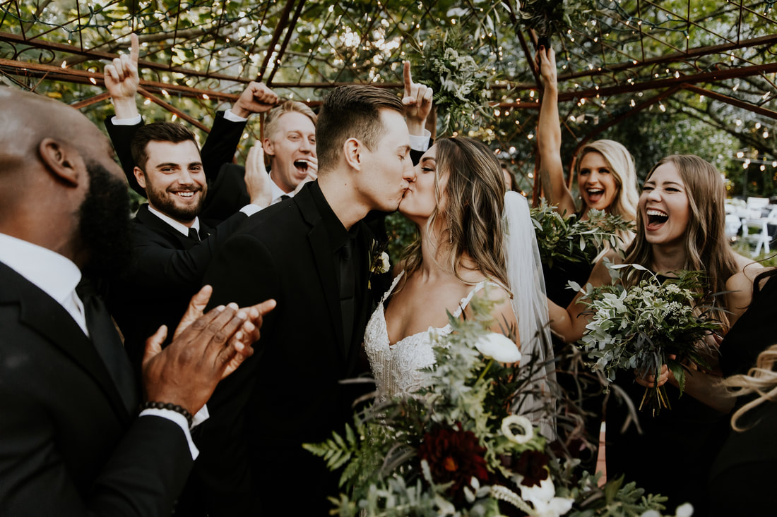wedding party in all-black cheering on newlyweds as they kiss in gazebo