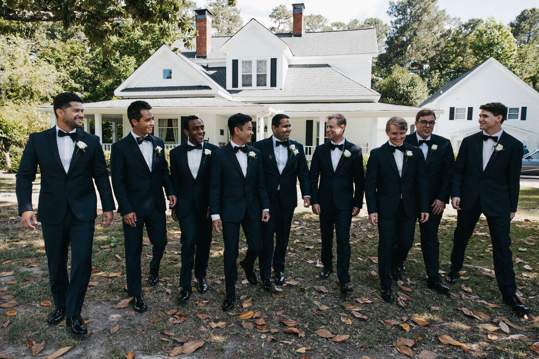 groom and groomsmen in black tuxes walking in front of white farmhouse