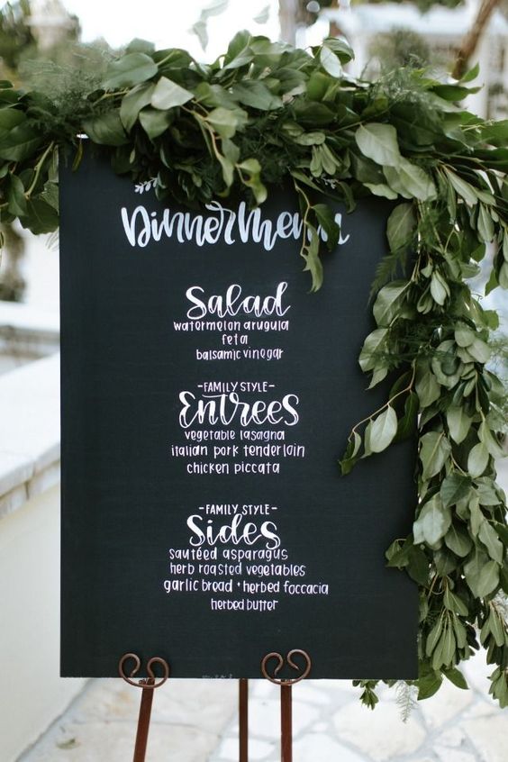 Black wedding menu chalkboard sign with large greenery hanging on top.