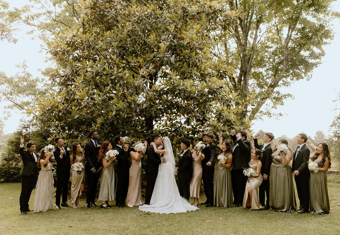 bride and groom kissing in front of tree while wedding party cheers