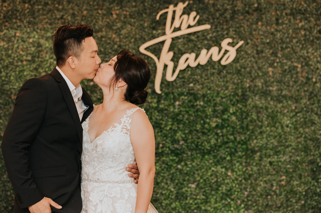 The couple kisses in front of a greenery wall photobooth backdrop with a wood sign of their last name.