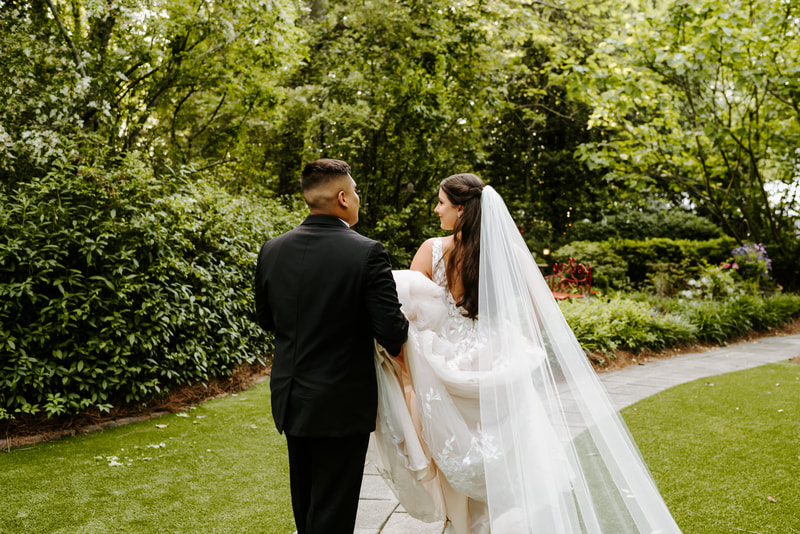 groom holding back of bride's dress as they walk through garden