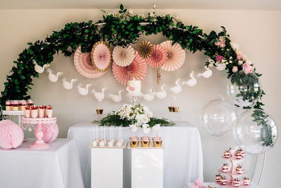 Baby shower decorated as Swan Lake with pink desserts and paper decorations, swan shaped banners, greenery, and pink and white flowers.